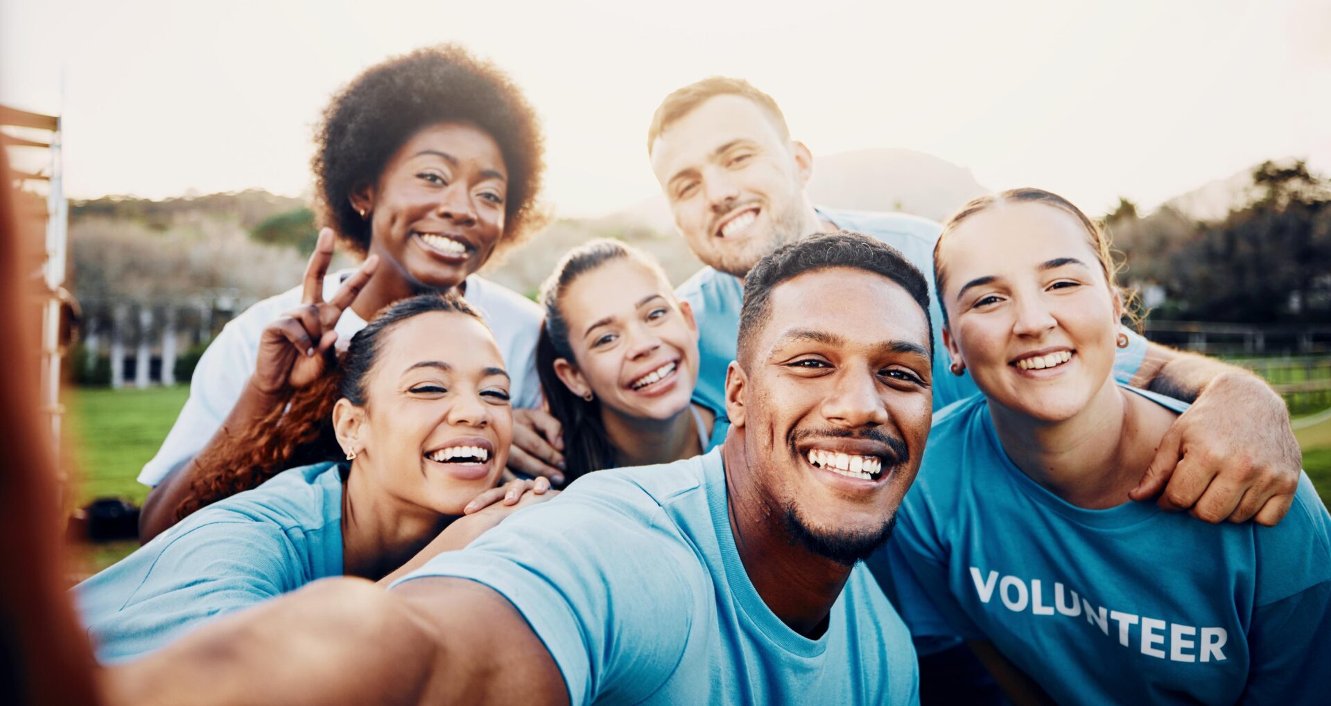 A diverse group of seven smiling people in light blue shirts, one with "VOLUNTEER" printed on it, pose for a group selfie outdoors.
