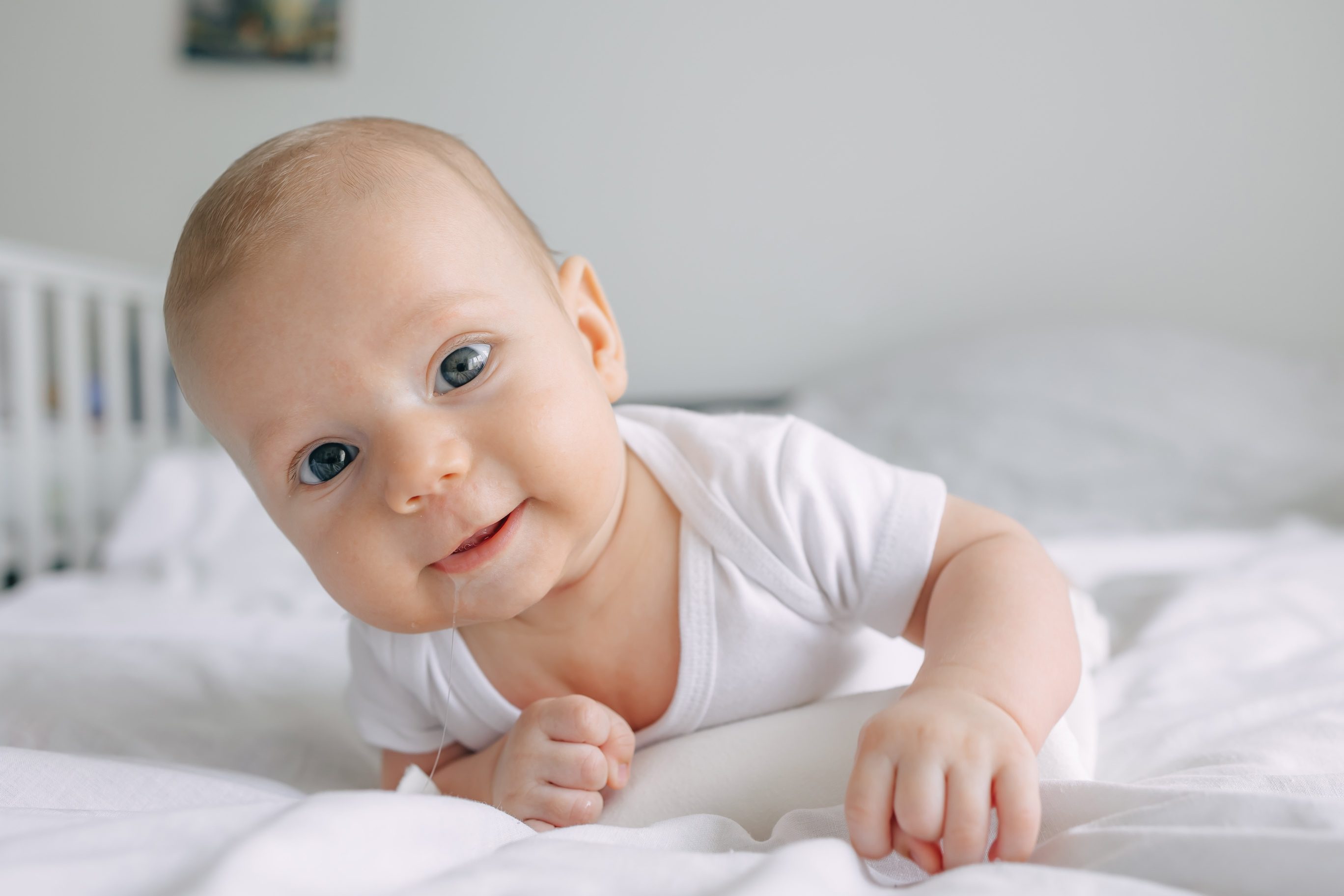 Baby in a white onesie lies on a bed, looking directly at the camera with a slight smile, surrounded by white bedding in a softly lit room.
